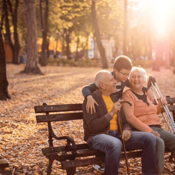Un couple de personnes âgées souriantes munis d'une canne et de béquilles est assis sur un banc en bois et est enlacé par un jeune homme.